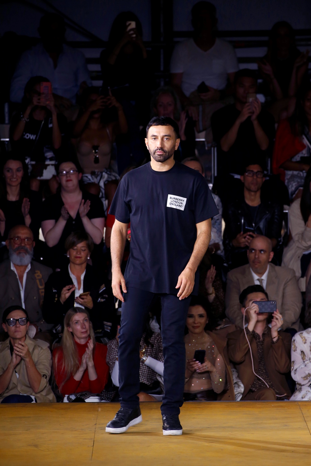 A imagem mostra o designer Riccardo Tisci andando em uma passarela com piso amarelo