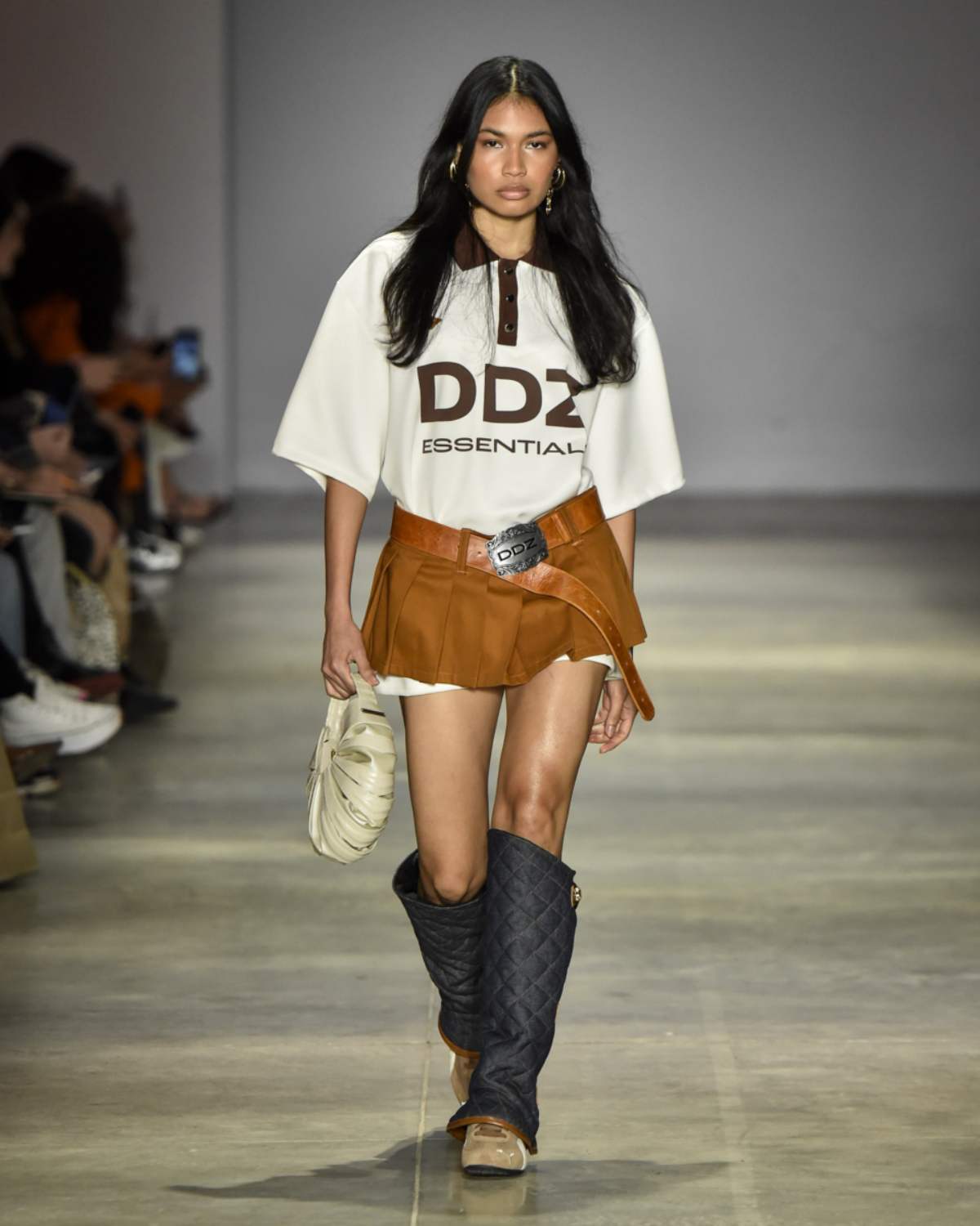 A imagem mostra uma mulher desfilando com uma camiseta branca com detalhes marrons, saia marrom, e bota preta