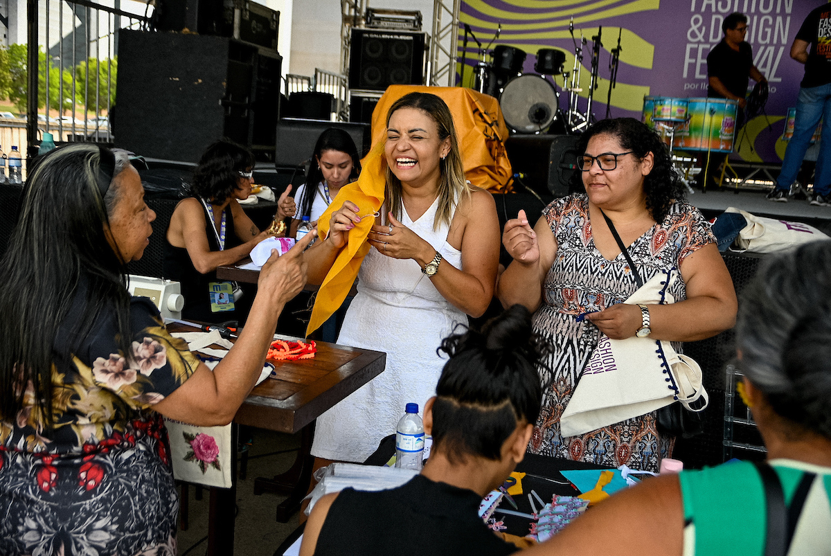 A imagem mostra uma mulher de vestido branco em pé falando com outras pessoas