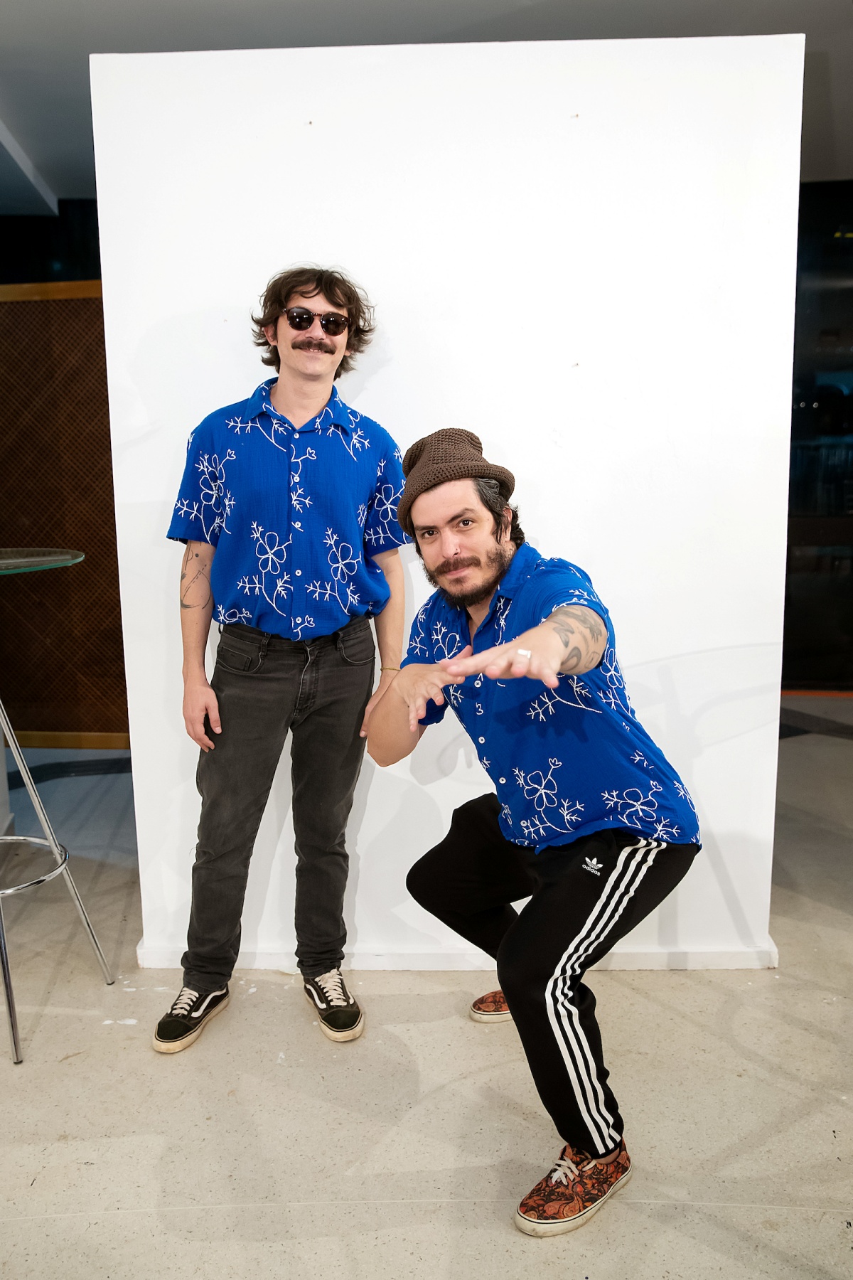 A imagem mostra dois homens combinando de camisa azul e calça preta fotografados em uma parede branca