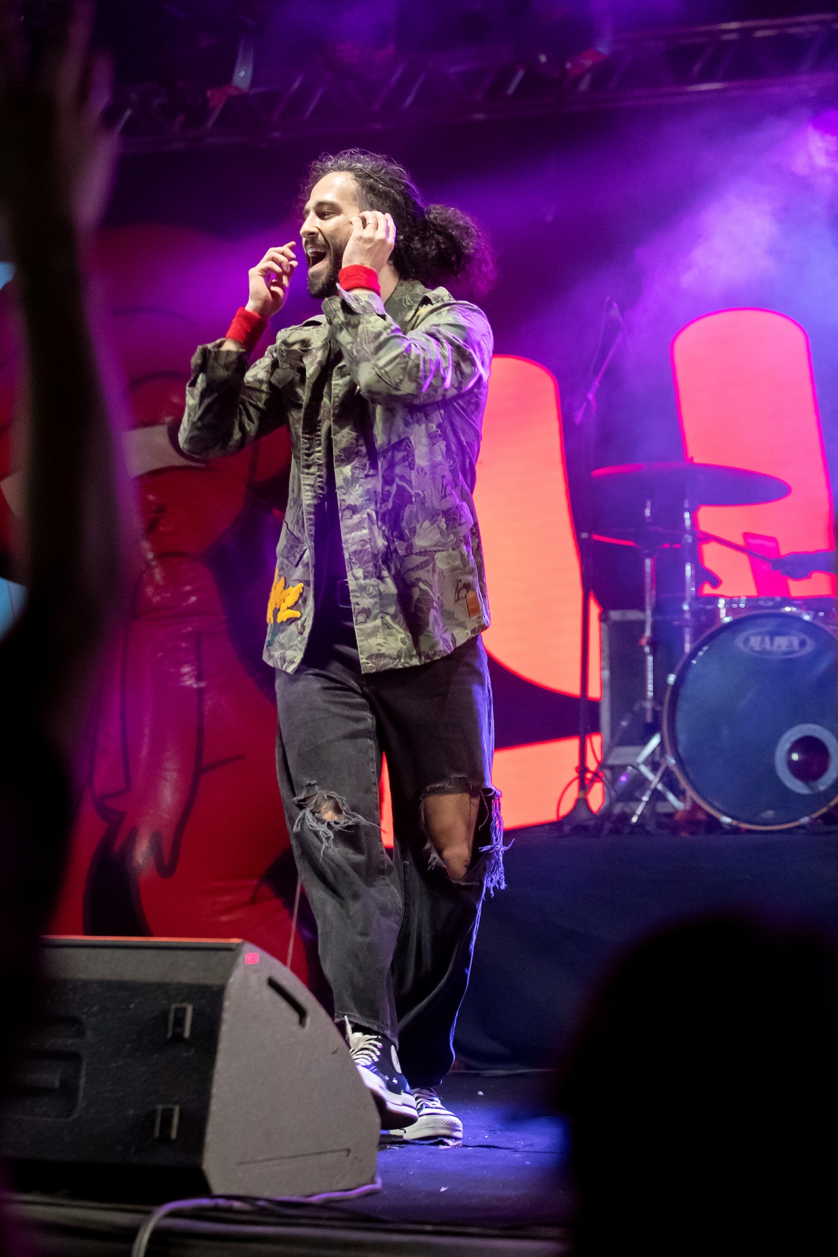 A imagem mostra um homem cantando em cima de um palco de jaqueta camuflada e calça jeans rasgada