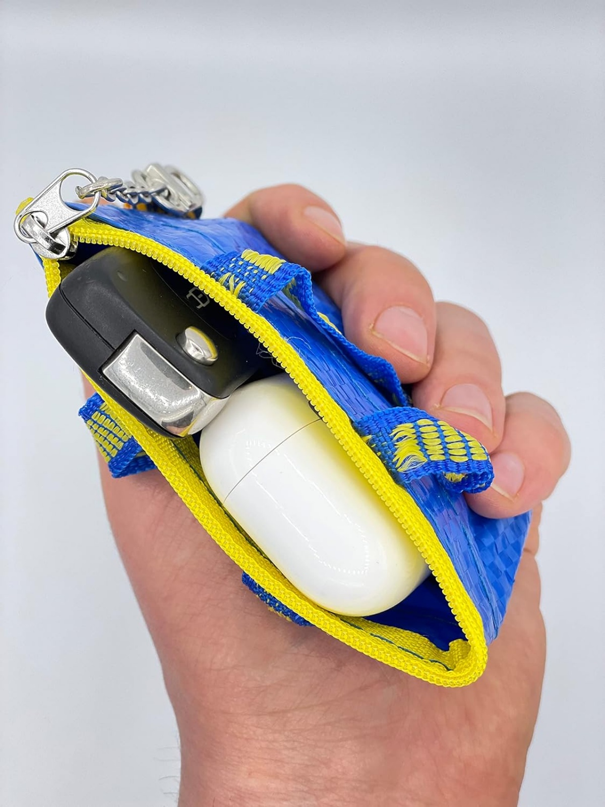 A image mostra uma mão carregando uma bolsinha azul com uma chave de carro e uma caixa de air pods dentro