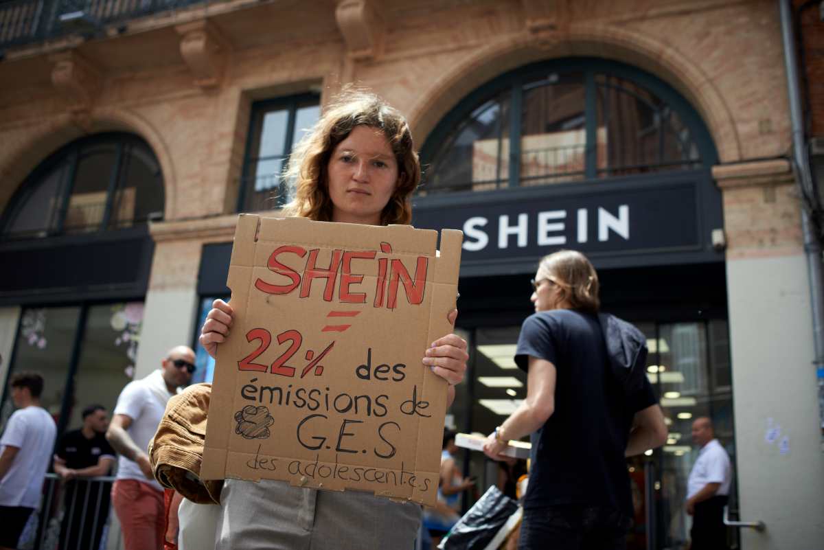 Uma jovem segura um cartaz com os dizeres "SHEIN = 22% do gás de aquecimento emitido para jovens" em frente a uma loja pop-up da SHEIN. 