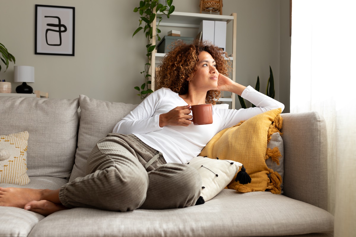 Imagem colorida de mulher descansando e tomando uma bebida no sofá - Metrópoles 