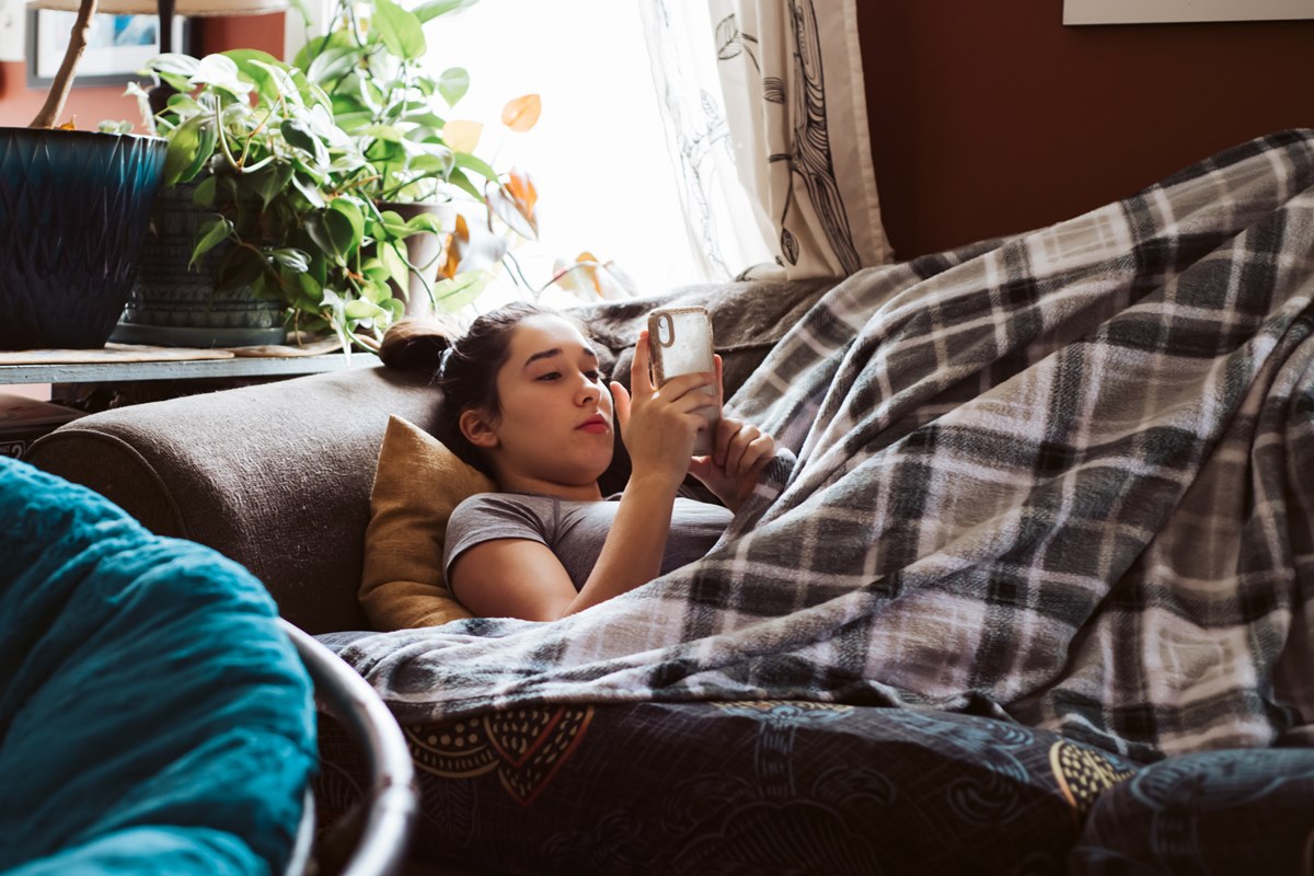 Adolescente deitada no sofá, debaixo de um cobertor, assistindo a algo no celular. Metrópoles