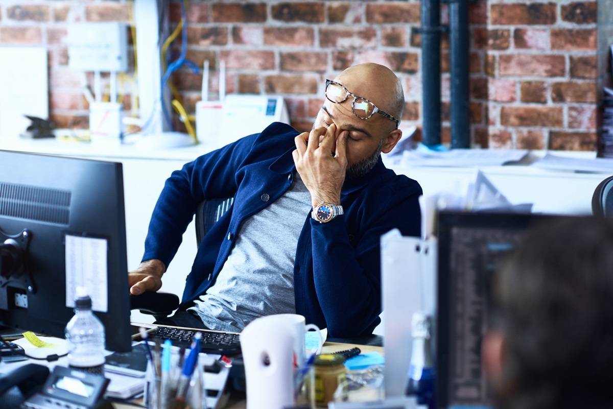 Homem cansado sentado à mesa em um escritório moderno. Metrópoles