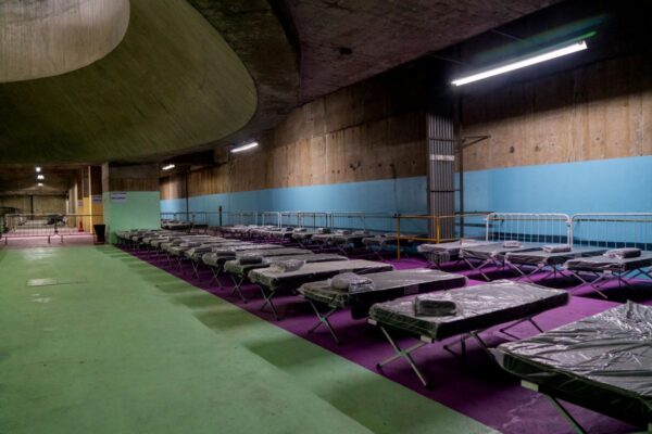 Imagens coloridas mostram várias camas improvisadas, com colchões e cobertores, lado a lado, sobre um tapete roxo, dentro de estação do metrô de São Paulo