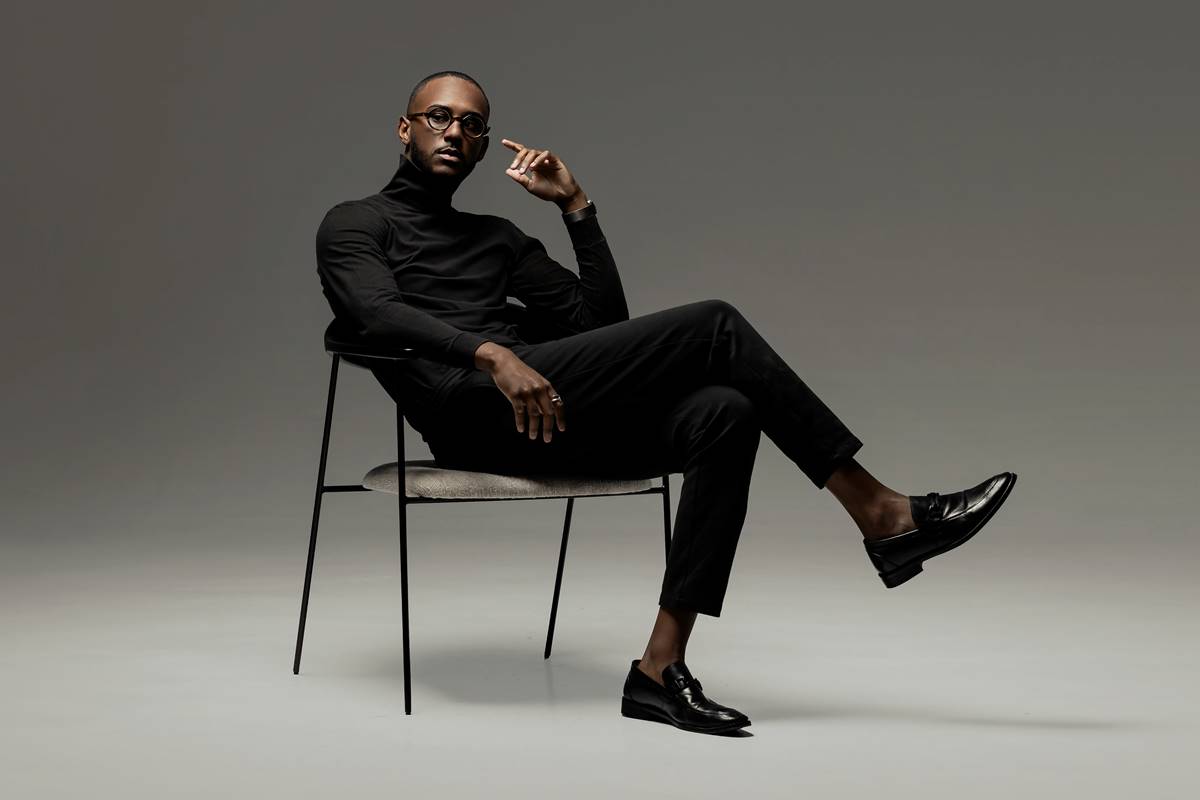 foto colorida de homem negro de blusa gola role preta e calça preta posando serio sentado em cadeira - metrópoles