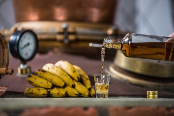 Foto colorida de cachaça sendo colocada em copo e ao lado uma penca de banana - Metrópoles