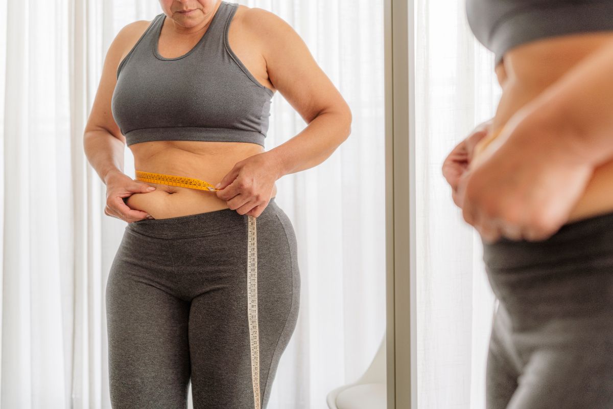 foto colorida mulher se olhando no espelho e pegando na gordura abdominal