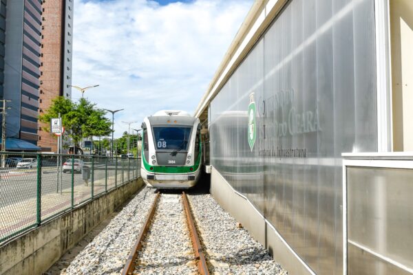 Trem verde se aproxima de plataforma ao lado de muro de vidro, com prédios altos ao fundo.