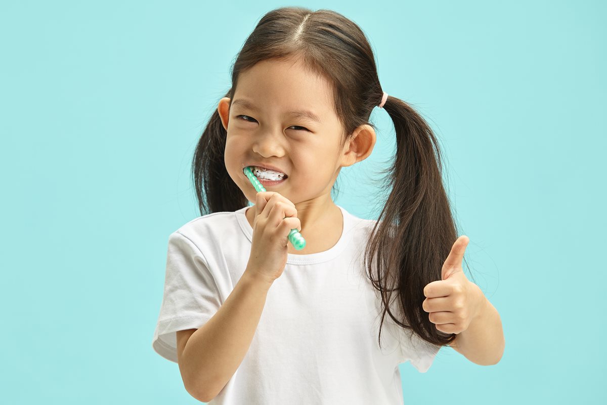 Foto colorida de criança fazendo sinal positivo com as mãos enquanto escova os dentes - Metrópoles.