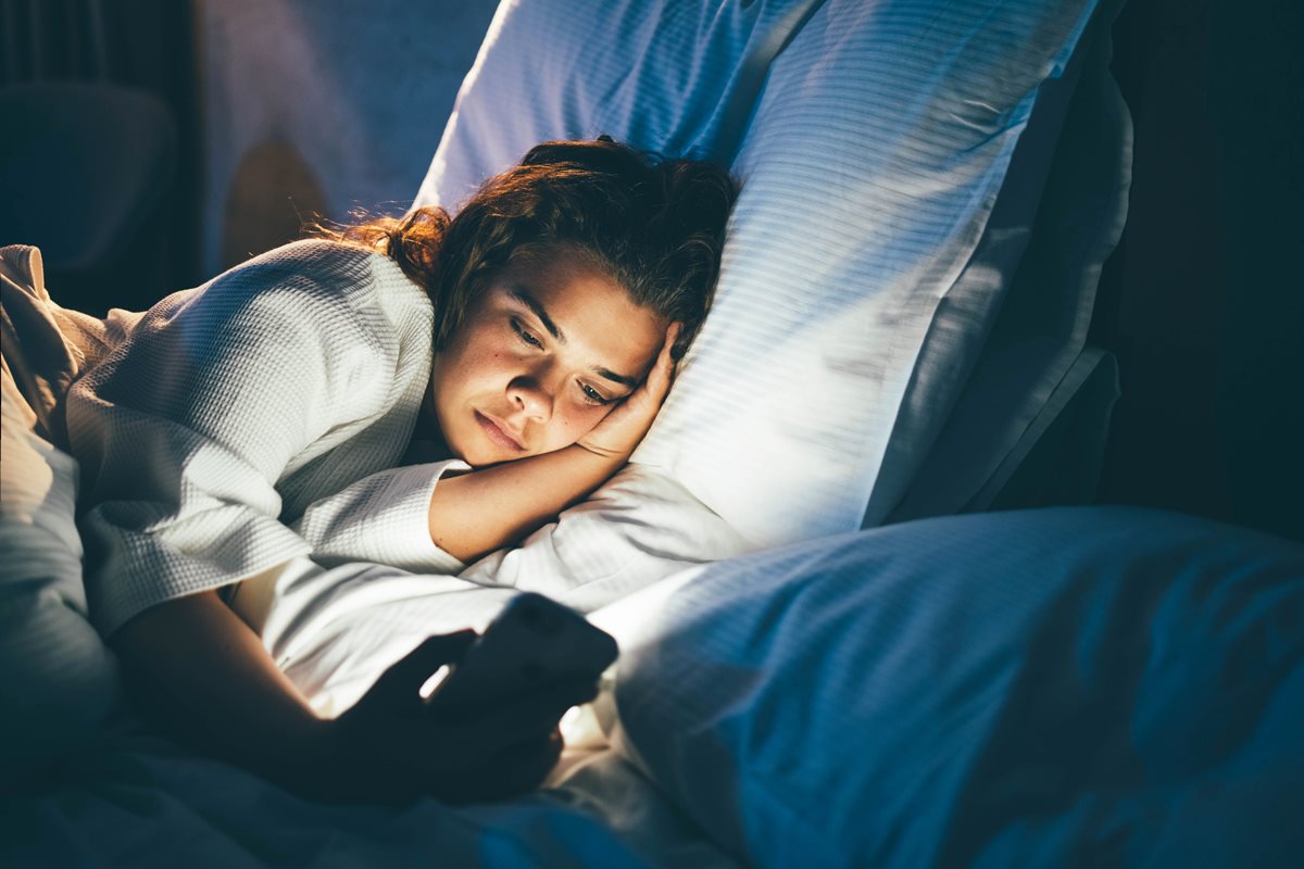 Foto colorida de mulher deitava sem conseguir dormir mexendo no celular - Metrópoles. 