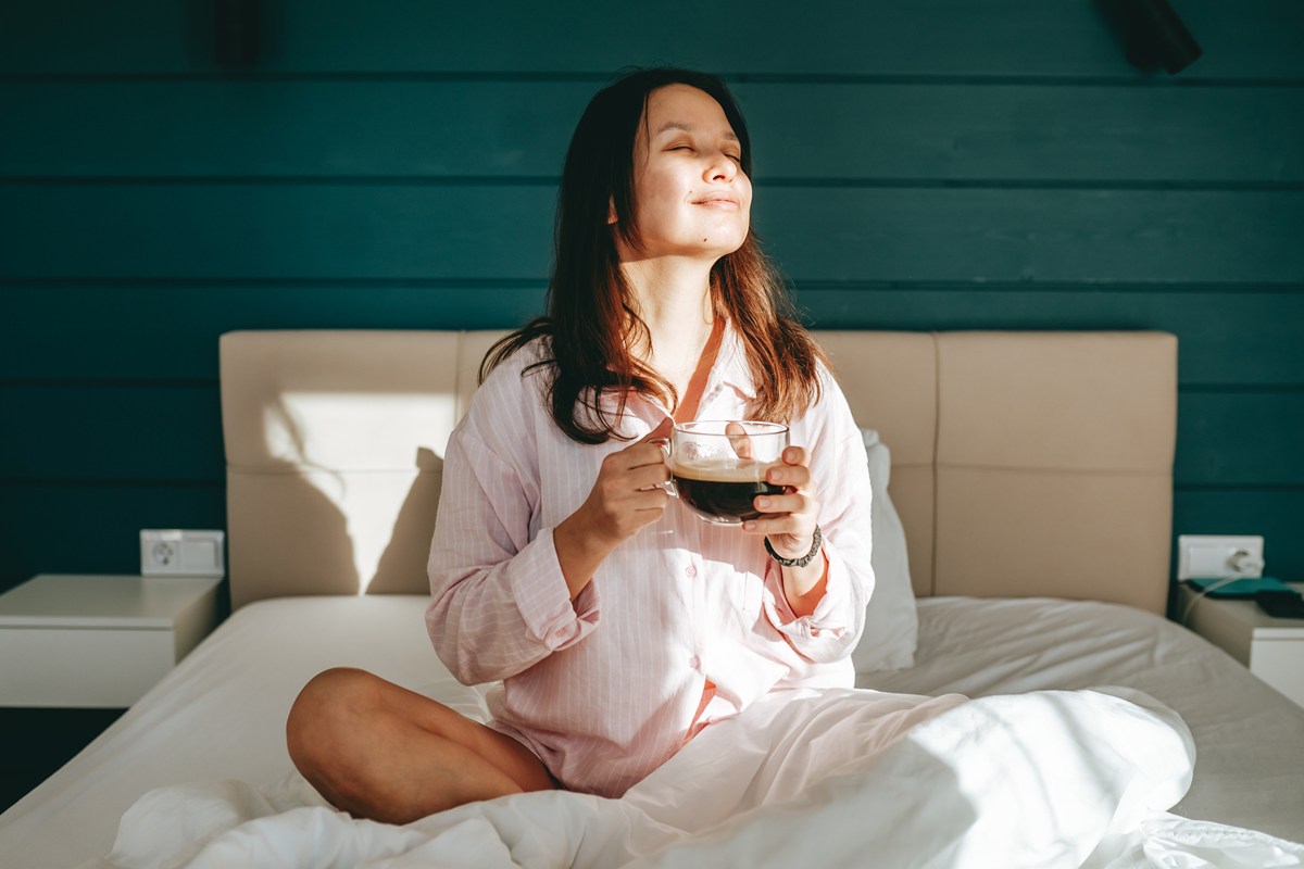 Foto colorida de mulher em cama segurando xícara de café - Metrópoles