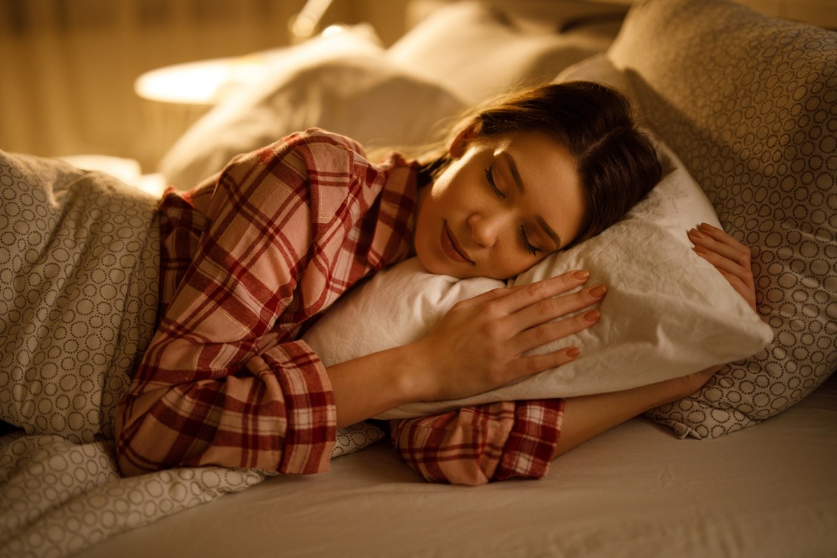 Mulher dormindo na cama abraçada a um travesseiro branco macio. Metrópoles