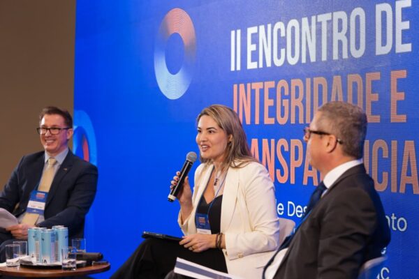 Auditório com plateia atenta a um painelista que fala no palco. Fundo azul com título do evento em destaque