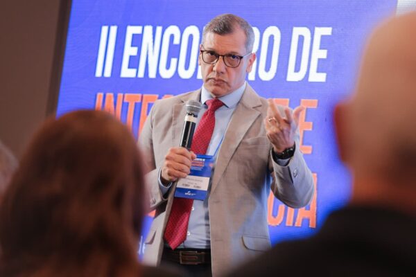 Homem de óculos e terno escuro fala em pé, gesticulando com a mão esquerda. O fundo azul traz o logotipo do Senac.