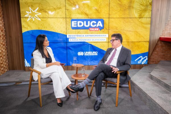 Uma mulher de blazer branco e calça clara, sentada em uma poltrona, conversa com um homem de terno escuro e gravata lilás. Eles estão frente a frente em um estúdio iluminado, com o fundo em tons amarelo e azul