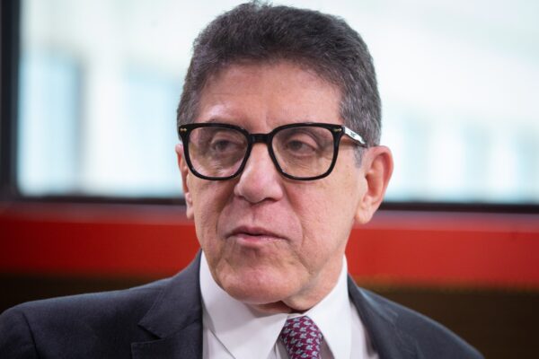 O rosto de David Uip aparece em primeiro plano. Ele usa óculos de armação preta, terno e gravata, com expressão séria e atenta