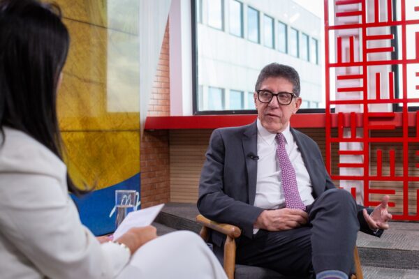 Cena de entrevista: a jornalista de branco segura um caderno e ouve atenta, enquanto o médico, de terno escuro, está sentado em frente a ela. O fundo mostra painéis coloridos em vermelho, azul e amarelo
