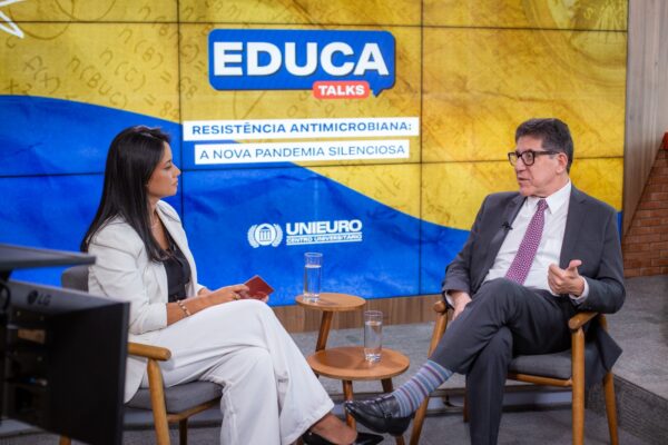 O médico aparece sentado em poltrona, de pernas cruzadas, gesticulando com a mão direita. O fundo é amarelo, em um estúdio bem iluminado