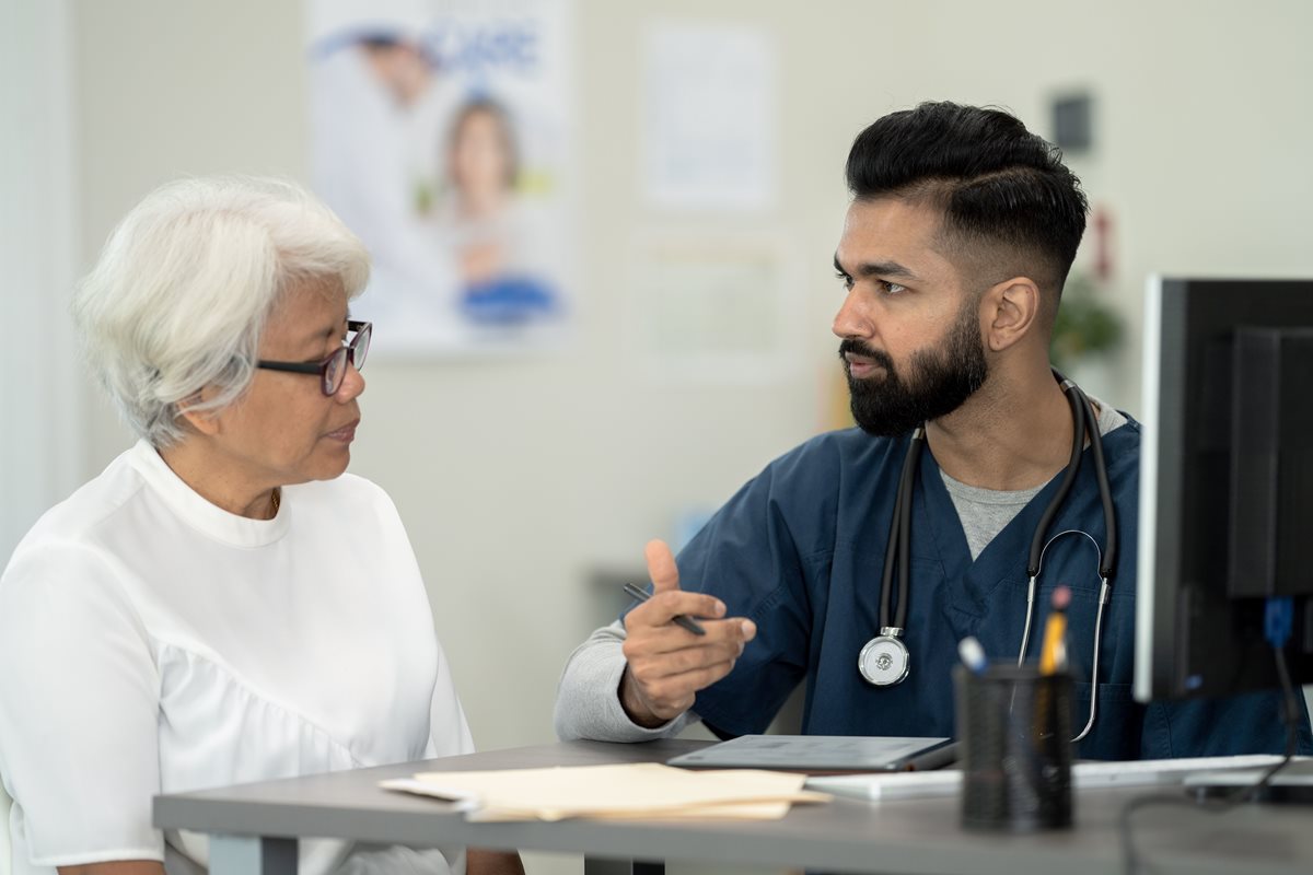 Foto colorida de um homem vestido de médico e uma senhora sendo atendida por ele em frente a um computdor - Metrópoles.
