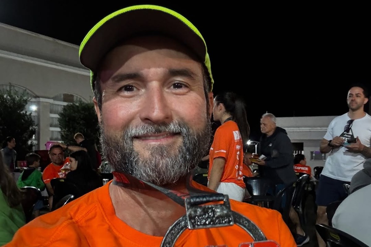 Foto colorida de homem sorrindo de boné e com blusa laranja. Ele segura medalha de corrida - Metrópoles