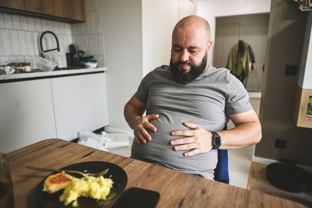 Foto colorida de homem careca e barba preta, em frente à mesa com as mãos na barriga olhando para o prato de comida. -metrópoles.