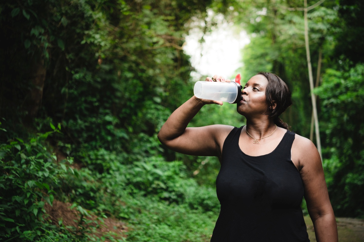 Foto colorida de mulher bebendo água em ambiente arborizado - Metrópoles.