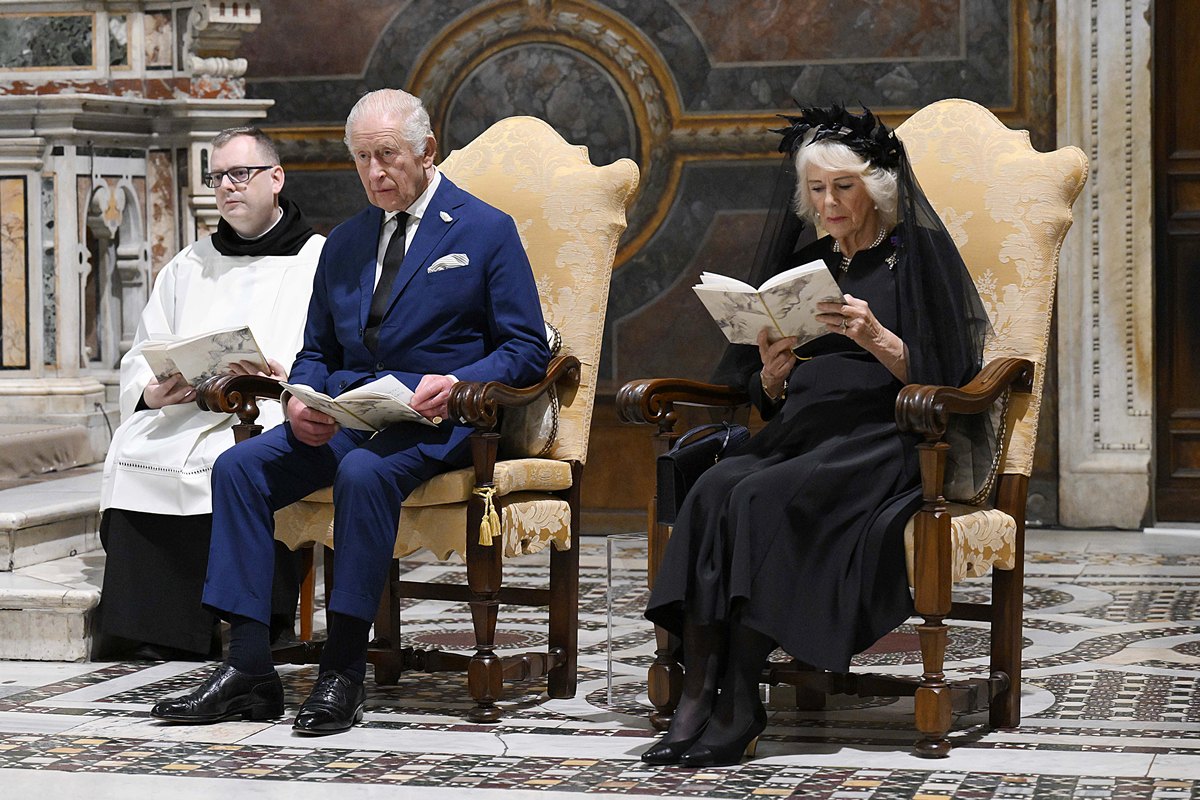 Foto colorida de homem com vestes religiosas ao lado de homem de terno e de mulher com roupas pretas. Eles acompanham uma cerimônia religiosa - Metrópoles