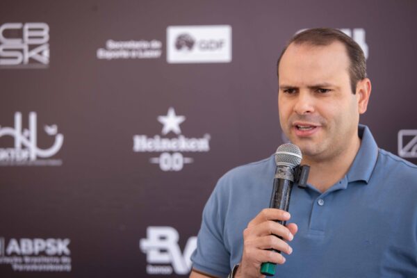 Homens com microfones falam durante entrevistas, alguns vestindo camisetas esportivas e crachás de identificação. O clima é descontraído