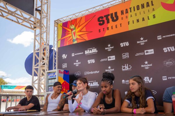 Um grupo de skatistas, homens e mulheres de diferentes idades e estilos, está reunido em uma mesa de coletiva, com microfones e backdrop do evento ao fundo. Alguns falam sorrindo, outros ouvem atentos. Ao fundo, o logotipo do STU National Street Finals 2025 e marcas de patrocinadores. A luz do dia ilumina o espaço montado em estrutura metálica.
