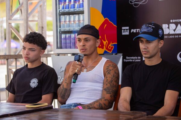 Um grupo de skatistas, homens e mulheres de diferentes idades e estilos, está reunido em uma mesa de coletiva, com microfones e backdrop do evento ao fundo. Alguns falam sorrindo, outros ouvem atentos. Ao fundo, o logotipo do STU National Street Finals 2025 e marcas de patrocinadores. A luz do dia ilumina o espaço montado em estrutura metálica.