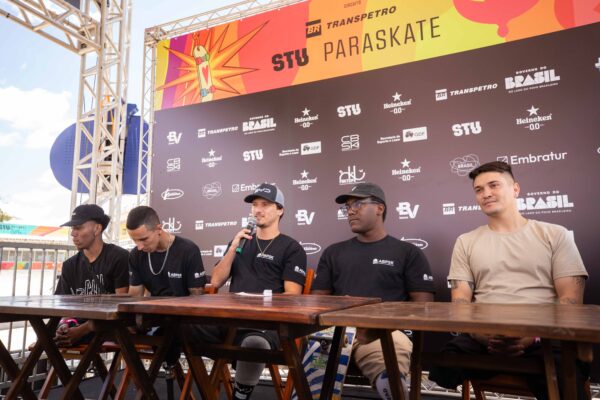 Homens com microfones falam durante entrevistas, alguns vestindo camisetas esportivas e crachás de identificação. O clima é descontraído e inspirador, com destaque para o sorriso dos atletas e o cenário ensolarado da pista ao fundo.