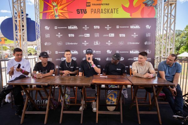 Homens com microfones falam durante entrevistas, alguns vestindo camisetas esportivas e crachás de identificação. O clima é descontraído e inspirador, com destaque para o sorriso dos atletas e o cenário ensolarado da pista ao fundo.