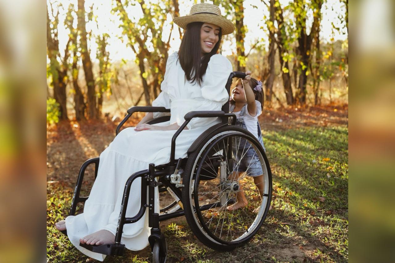Foto colorida de mulher jovem vestida de branco em uma cadeira de rodas enquato uma criança a empurra - Metrópoles. 