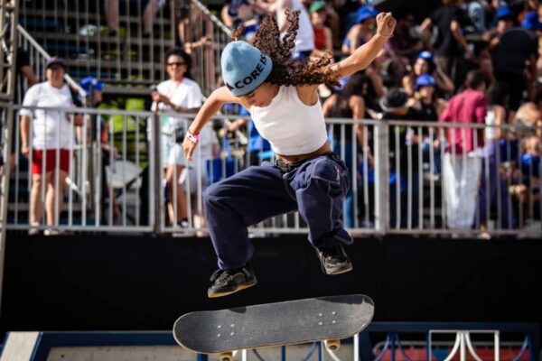 Um skatista de camiseta branca e calça larga realiza um salto sobre o skate em pleno ar, diante das arquibancadas cheias e sob o sol da tarde.