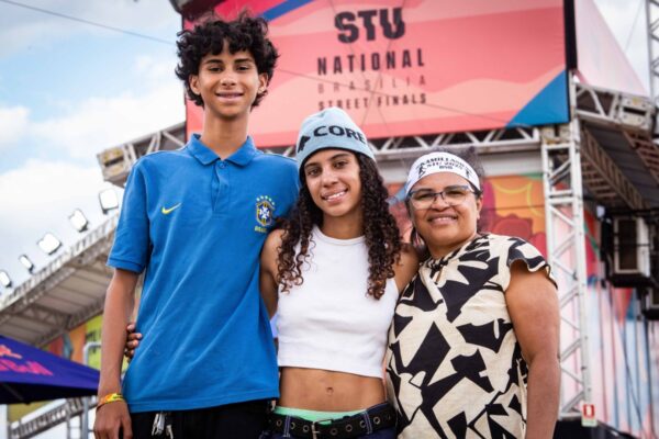 Três pessoas sorriem diante do painel do STU. A jovem atleta usa blusa branca e calça larga, abraçada ao irmão e à mãe, que veste camisa estampada.