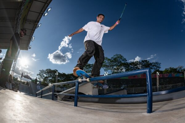 Um homem de camiseta clara e calça estampada desliza sobre o corrimão com o skate, sob céu azul e refletores da pista. Ele usa capacete branco com uma frase escrita à mão