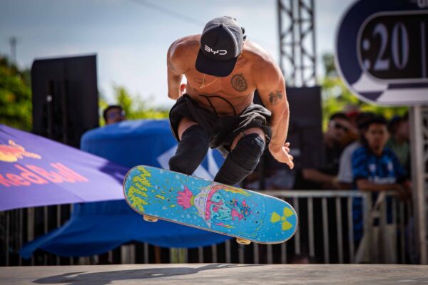Homem de boné e camiseta preta equilibra o skate sobre um corrimão prateado, sob o sol
