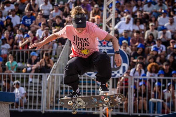 Uma skatista de camiseta rosa desliza sobre um corrimão com o skate, cercada por grades e espectadores atentos.