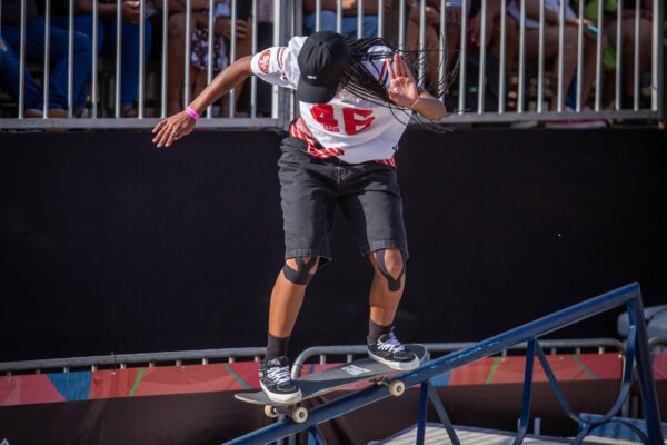 Uma jovem skatista faz uma manobra sobre o corrimão da pista de skate sob o sol, usando calça preta e camiseta clara, enquanto o público assiste atrás das grades.