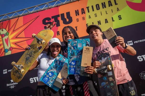 Três skatistas posam sorrindo no pódio colorido, segurando troféus e medalhas, com o letreiro “STU National Finals” ao fundo.