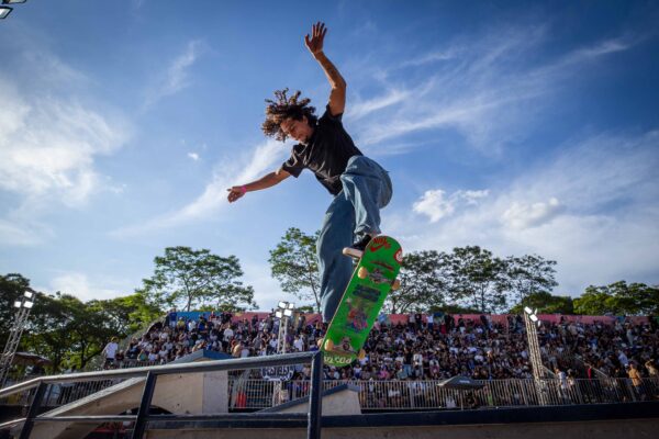 Vinicius Costa, 24 anos, é captado no ar durante uma manobra sobre um obstáculo metálico: o corpo inclinado para a frente, joelhos dobrados, braços equilibrando o movimento. Ele usa camiseta escura e jeans; o shape do skate é verde. Ao fundo, a arquibancada lotada e o céu azul, com luz de fim de tarde iluminando a cena