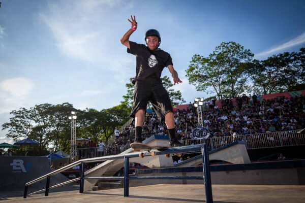 Um skatista em movimento salta com o skate sobre um obstáculo, capturado no ar sob luz intensa, com expressão concentrada e o público ao fundo.