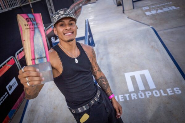 Um skatista sorri segurando o troféu do circuito com o logo “Metrópoles” no chão da pista, vestindo regata preta e boné, com tatuagens visíveis no braço