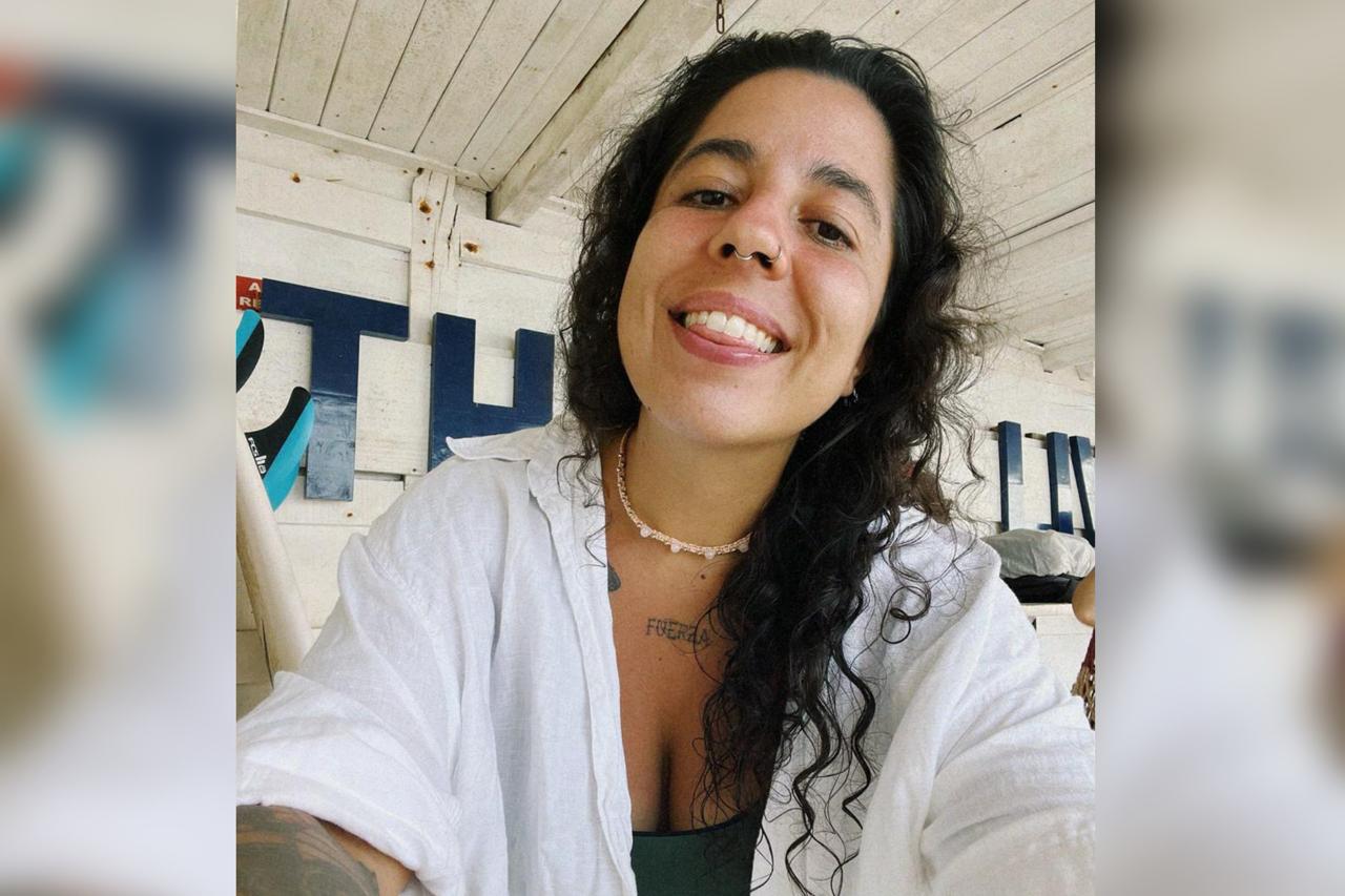 Foto colorida de mulher jovem que sorri para foto vestida com blusa branca - Metrópoles. 