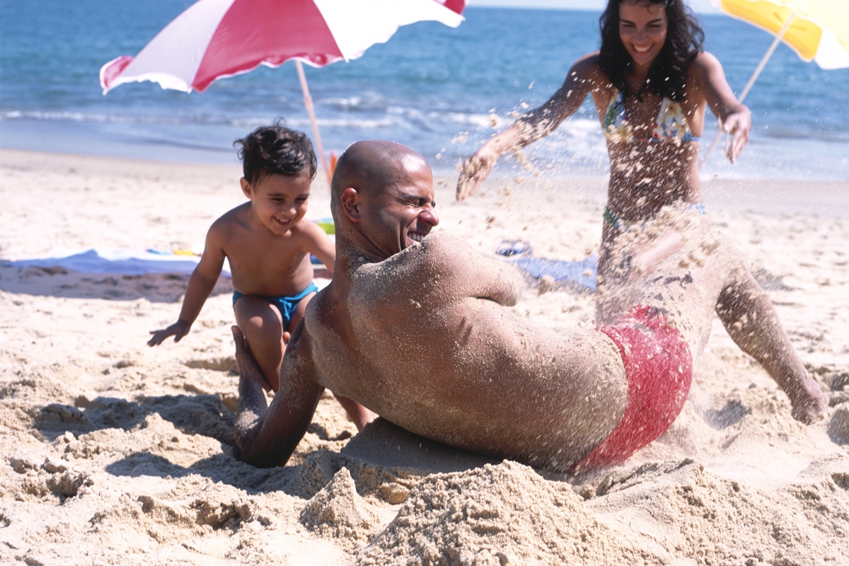 Imagem colorida mostra homem negro careca brincando na praia com uma criança e uma mulher brancas - Metrópoles 