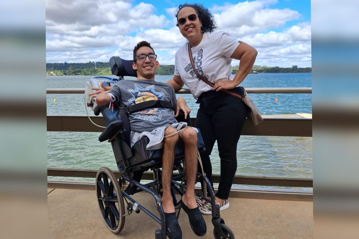 Foto colorida de mulher ao lado de jovem cadeirante, em frente a um lado - Metrópoles.